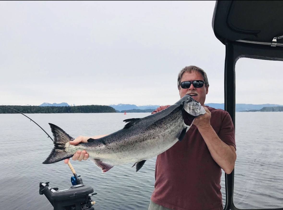 A man holding a large fish he caught, he is smiling and wearing a red shirt and sunglasses. Mountains, calm waters, the treeline and cloudy skies are visible in the background.